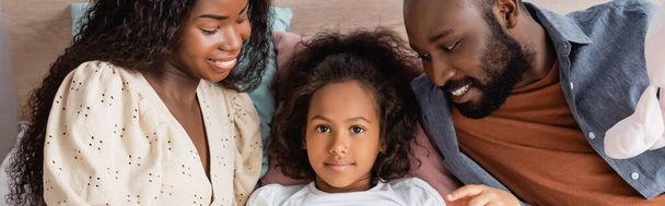 panoramic crop of african american child lying in bed and looking at camera near parents - Фото, изображение