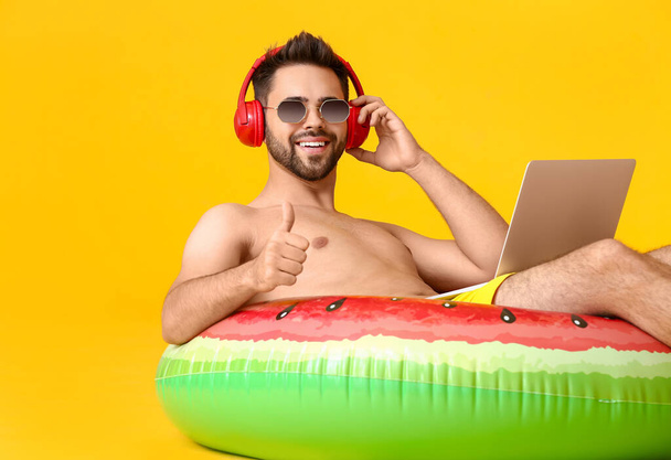 Jongeman met koptelefoon, opblaasbare ring en laptop met duim-up gebaar op kleur achtergrond - Foto, afbeelding