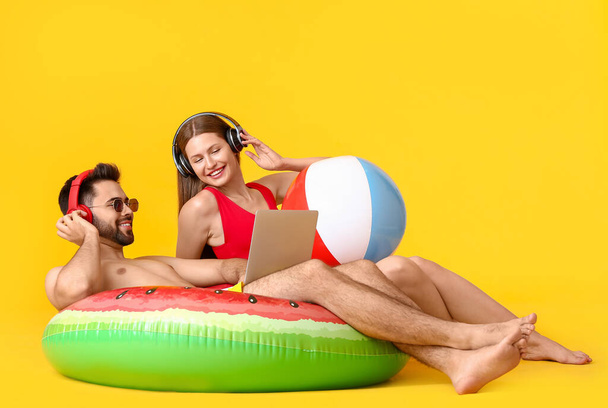 Young couple with headphones, inflatable ring and laptop on color background - Photo, Image