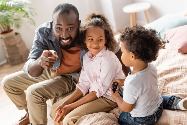 excited african american man pointing with finger near children sitting on bed - Photo, Image