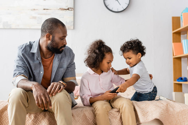 афроамериканський чоловік у повсякденному одязі сидить на ліжку біля дітей тримає телевізор пульт дистанційного керування
  - Фото, зображення