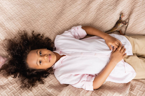 vue du dessus de la fille afro-américaine en chemise blanche reposant dans le lit tout en regardant la caméra - Photo, image