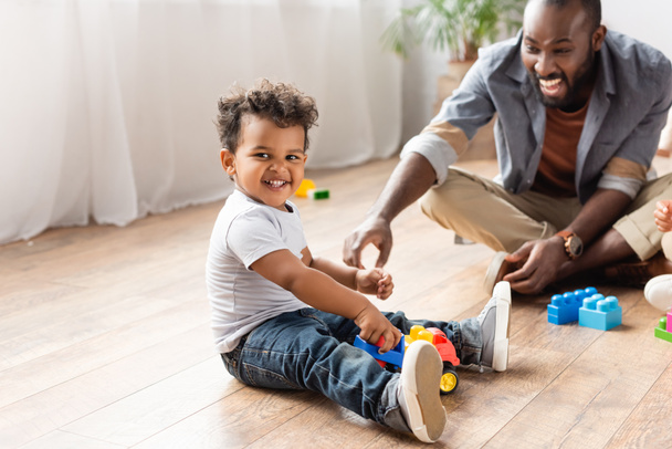 animado afro-americano homem perto filho brincando com brinquedo caminhão no chão de madeira - Foto, Imagem