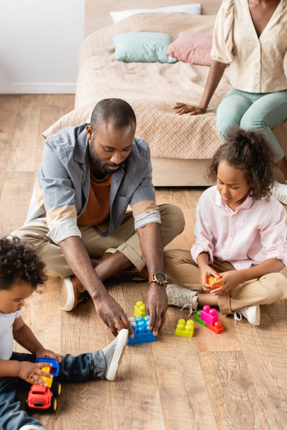 high angle view of african american man with daughter and son playing with building blocks on floor near mother sitting on bed - Fotó, kép