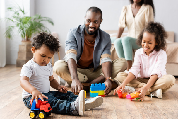 selectieve focus van Afrikaans-Amerikaanse man en kinderen spelen met bouwstenen op de vloer in de buurt van moeder zitten op bed - Foto, afbeelding