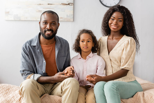 african american husband and wife touching hands of daughter while sitting in bedroom - Photo, Image