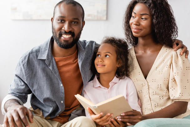 young african american man touching shoulder of wife while sitting near daughter holding book - Foto, imagen