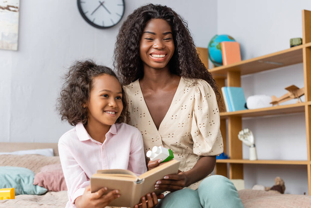 african american girl holding book while sitting in bedroom with mother - 写真・画像