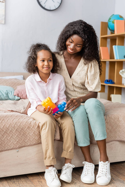 african american girl holding building blocks while sitting on bed near mother - Foto, afbeelding
