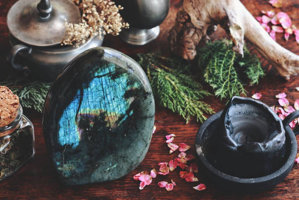 Labradorite crystal on dark wooden table with various nature objects like dried evergreens, plants, flowers, herbs in the background, Messy and cluttered wiccan witch altar with big labradorite on it - Photo, Image