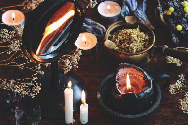 Candle flame divination in concave mirror on wiccan witch altar. Black candle burning on a messy cluttered table with dried herbs and flowers, crystals, plants, other nature objects in dark background - Photo, Image