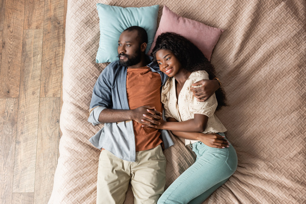 bovenaanzicht van afrikaanse amerikaanse man en vrouw hand in hand en kijken weg terwijl liggend in bed in casual kleding - Foto, afbeelding