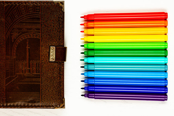 коричневый кожаный дневник и яркие цветные маркеры без крышек лежат на белом деревянном столе - Фото, изображение