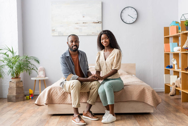 fiatal afro-amerikai pár alkalmi ruhák fogják egymás kezét, és nézi a kamera, miközben ül az ágyban - Fotó, kép