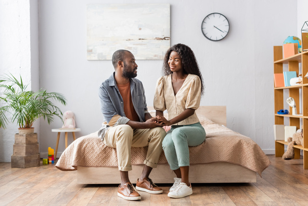 joven africano americano pareja en casual ropa cogido de la mano y mirando uno al otro en dormitorio - Foto, Imagen
