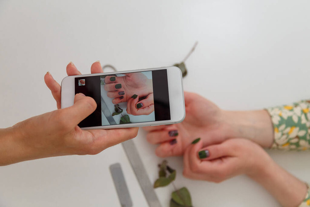 Nail artist taking photo with smartphone of her work, clients manicured hands. Green nail polish design, nail files and eucalyptus branch on white table. Spring manicure. Copy space. - Photo, Image