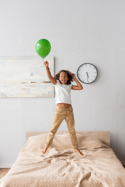 excited african american girl in casual clothes jumping on bed with red balloon  - Фото, изображение