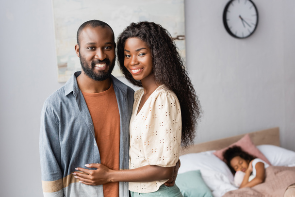 joven africano americano pareja mirando cámara mientras de pie cerca hija durmiendo en cama - Foto, imagen
