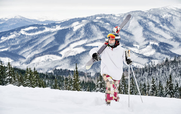Οριζόντια στιγμιότυπο του νεαρού σκιέρ σε πολύχρωμο κοστούμι περπάτημα κατά μήκος χιονισμένο λόφο, carring σκι του, κατά τη διάρκεια εκπληκτικά βουνά στο παρασκήνιο. Εμπρόσθια όψη, χώρος αντιγραφής, πλήρες μήκος - Φωτογραφία, εικόνα