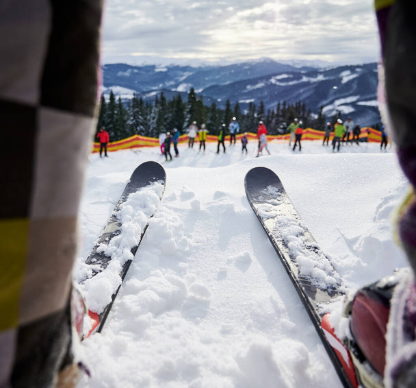 Primer plano del hombre en esquís de pie sobre nieve blanca profunda en la estación de esquí. Esquís alpinos unidos a botas de esquí con fijaciones de esquí y grupo de personas con montañas sobre fondo borroso. Concepto de esquí - Foto, Imagen