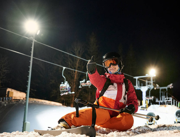 Girl skier wearing vivid suit sitting on snow-covered ski slope under illuminated chairlift at night, showing thumb up happy and satisfied after freeride at ski resort. Concept of extreme sport - Photo, Image