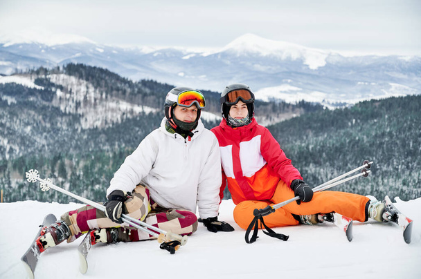 Two friends wearing ski suits and ski equipment are in winter mountains enjoying snowy weather, skiing and incredible landscapes. Portrait of a couple sitting on snowy hill smiling looking at camera - Photo, Image