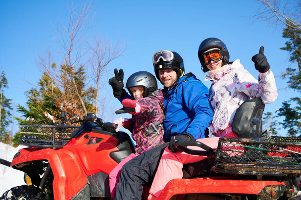 Young man and woman showing victory gesture and thumbs up while sitting on quad bike with adorable daughter. Girl sitting on all-terrain vehicle with parents. Concept of family and active leisure. - Photo, Image