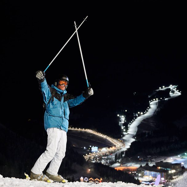 Esquiador masculino en chaqueta de esquí de invierno y casco levantando bastones de esquí y mirando a la cámara. Joven con gafas de esquí de pie en la pista cubierta de nieve con estación de esquí nocturno en el fondo. Concepto de esquí nocturno - Foto, Imagen