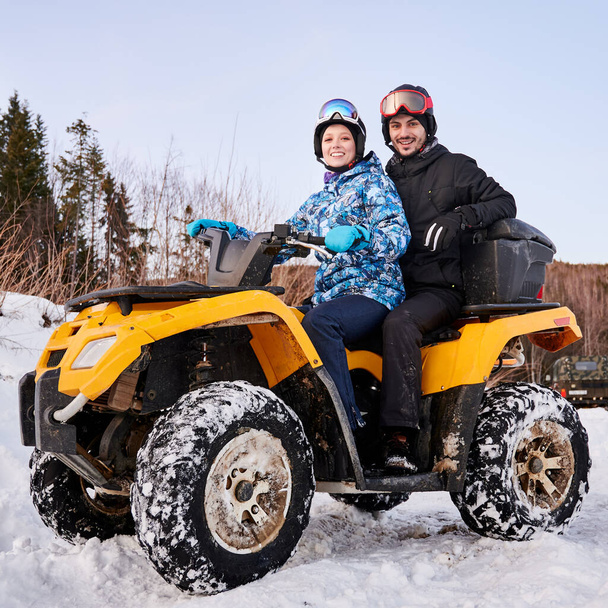 Portrait of romantic couple outdoor in winter in wild nature. Woman and man on yellow four-wheeler ATV in snowy mountains smiling at camera. Concept of relationships, active leisure, winter activities - Photo, Image