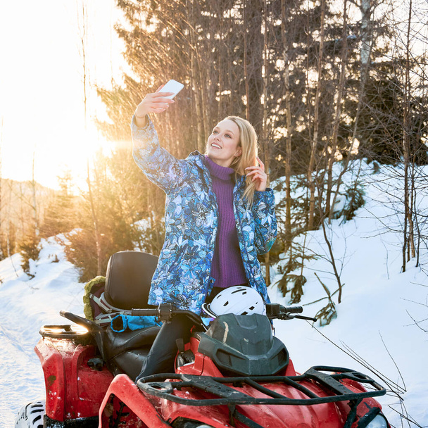 Mujer rubia alegre en chaqueta de invierno tomando fotos con teléfono inteligente durante el ciclismo cuádruple en las montañas de invierno. Encantadora viajera femenina en vehículo todo terreno mirando la cámara del teléfono y sonriendo. - Foto, Imagen