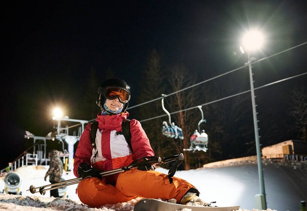 Esquiadora vistiendo un traje vívido, sentada en una pista de esquí cubierta de nieve, feliz y satisfecha después de freeride. Paseo nocturno en la estación de esquí. Concepto de esquí nocturno, actividades deportivas de invierno. - Foto, Imagen