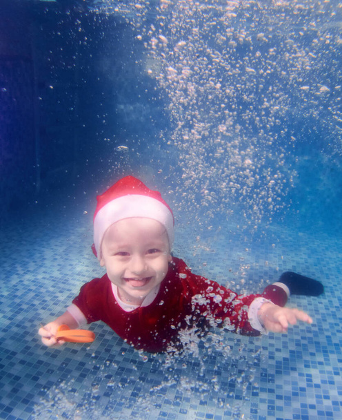 Front view of adorable baby boy in Christmas costume learning to swim underwater. Cute child balancing in crystal blue water and smiling. Concept of happy childhood, holiday and swimming. - Photo, Image