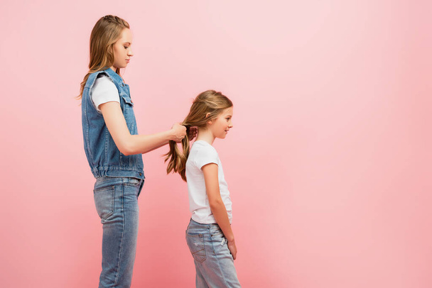 side view of young woman in denim clothes braiding hair of daughter isolated on pink - Фото, изображение