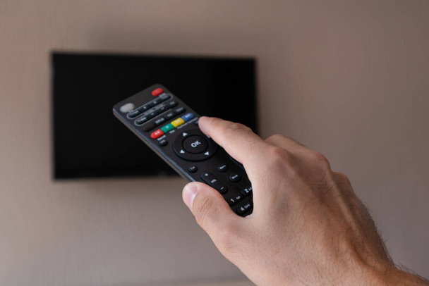 Mão masculina com controle remoto de tv e televisão borrada com uma tela preta no fundo. O homem muda de canal na TV. Espaço de cópia para design, close-up, foco seletivo
 - Foto, Imagem
