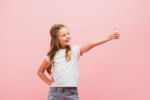 girl in white t-shirt with hand on hip looking away while showing thumb up isolated on pink - Fotografie, Obrázek