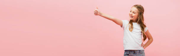 horizontal image of girl in white t-shirt with hand on hip showing thumb up while looking away isolated on pink - Photo, Image