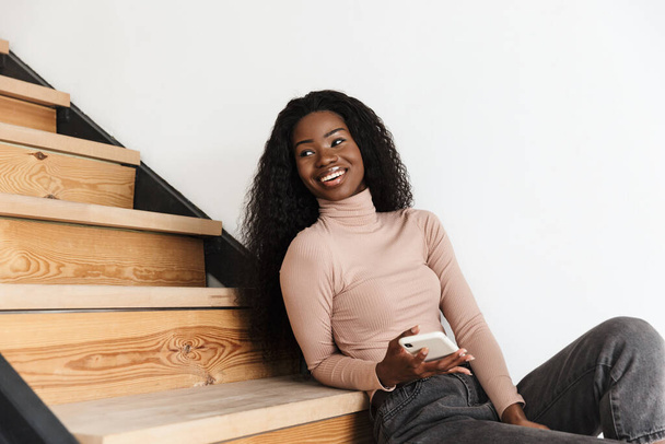 Image of beautiful happy positive cute young african woman sitting on stairs indoors at home while using mobile phone - Foto, Bild
