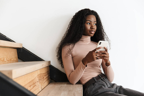 Image of beautiful concentrated young african woman sitting on stairs indoors at home while using mobile phone - Foto, imagen