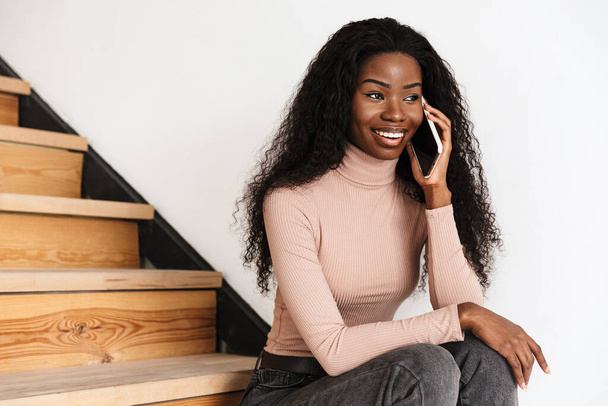 Image of pretty happy young african woman sitting on stairs indoors at home while talking by mobile phone - Φωτογραφία, εικόνα