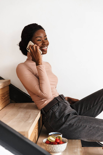 Image of pretty happy positive young african woman sitting on stairs indoors at home while talking by mobile phone - Photo, image
