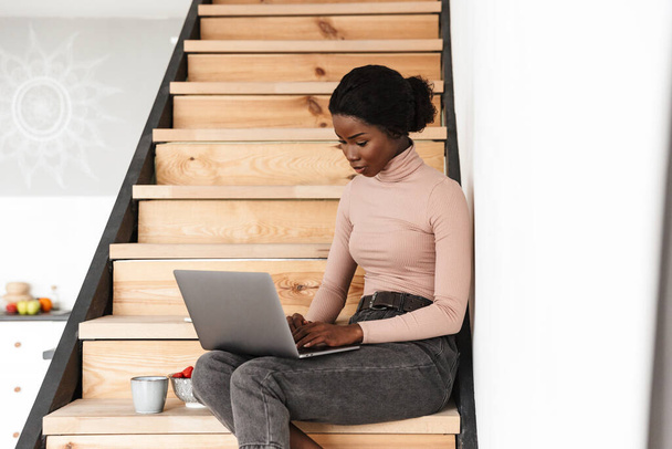 Image of beautiful concentrated young african woman sitting on stairs indoors at home while using laptop computer - Photo, image