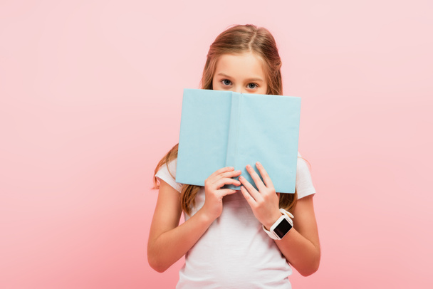 niño en smartwatch oscurecer la cara con libro mientras mira la cámara aislada en rosa - Foto, Imagen