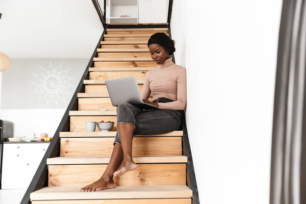 Image of beautiful concentrated young african woman sitting on stairs indoors at home while using laptop computer - Foto, Imagen