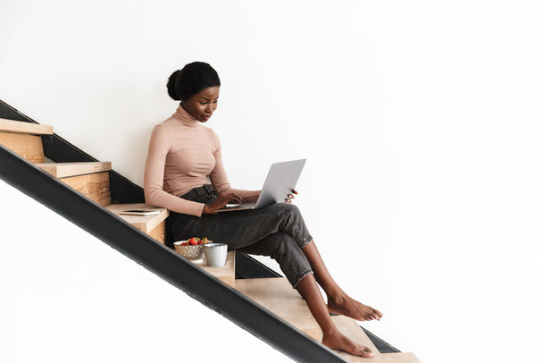 Jovem mulher africana atraente sentada em uma escada em casa, trabalhando no computador portátil, tomando café da manhã
 - Foto, Imagem