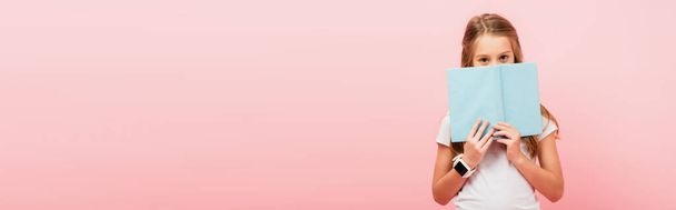 horizontales Bild von Mädchen in Smartwatch verschleiert Gesicht mit Buch, während Blick auf Kamera isoliert auf rosa - Foto, Bild