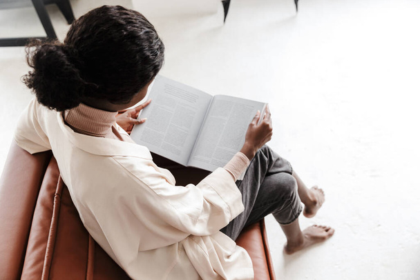 Vue arrière de la jeune femme africaine assise sur un canapé à l'intérieur de la maison en lisant un magazine ou un livre - Photo, image