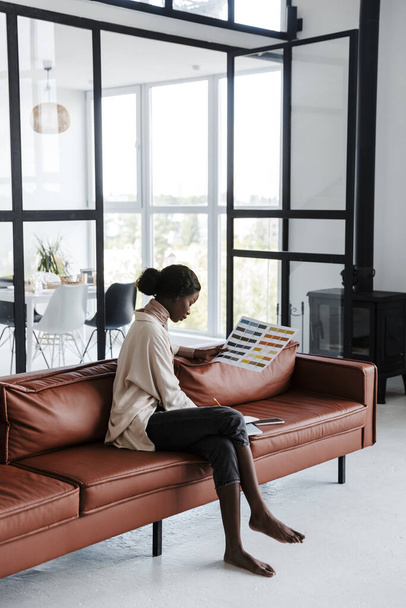 Photo of young african concentrated woman interior designer sitting on sofa indoors at home while working with catalogue of colors - Foto, immagini