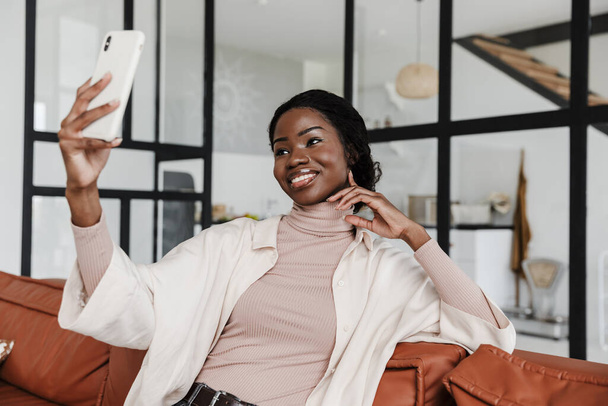 Image of amazing cheerful positive young african woman sitting on sofa indoors at home while take a selfie by mobile phone - Zdjęcie, obraz