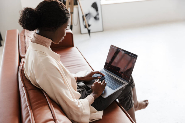 Side view image of young african concentrated woman sitting on sofa indoors at home while working with laptop computer - Foto, Imagem