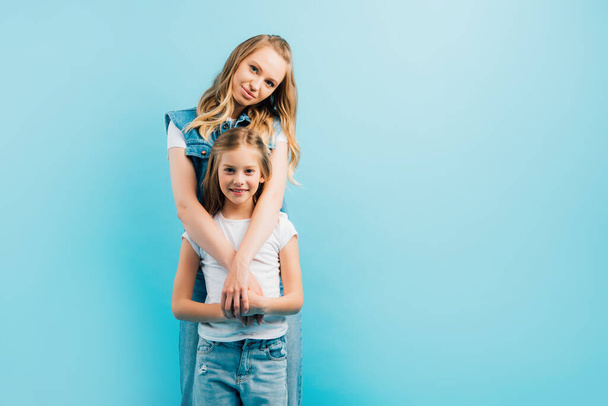 mother embracing child wearing white t-shirt and jeans while looking at camera isolated on blue - Photo, Image